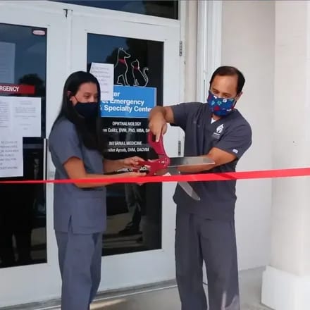 Two people in grey scrubs and masks cutting a red ribbon in front of a glass door with a sign