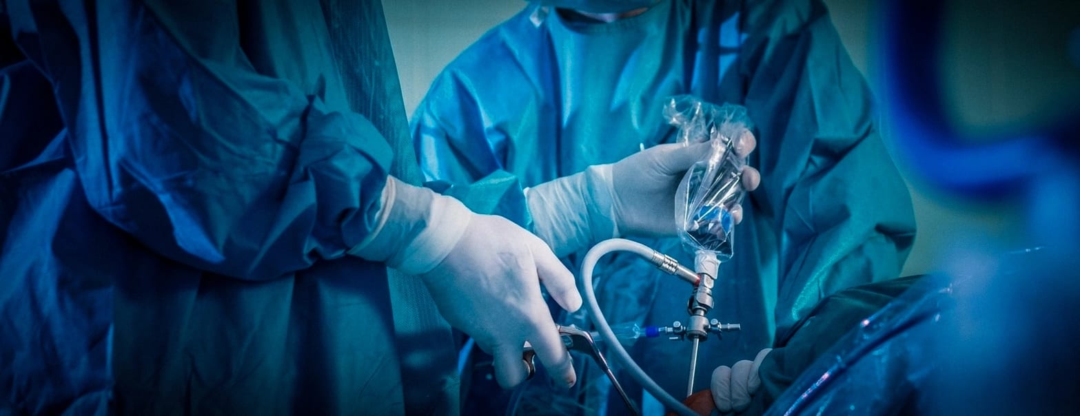 Gloved hands in blue surgical gowns holding a metal instrument attached to a clear tube